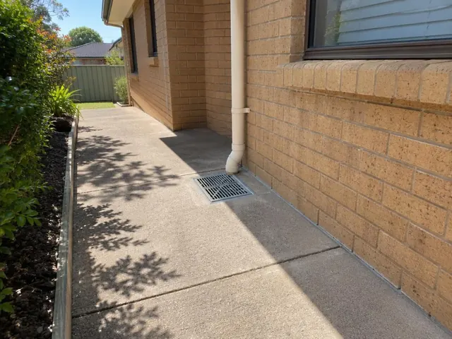 Outdoor gully grate beside a brick house wall on a dry concrete path in Lalor Park