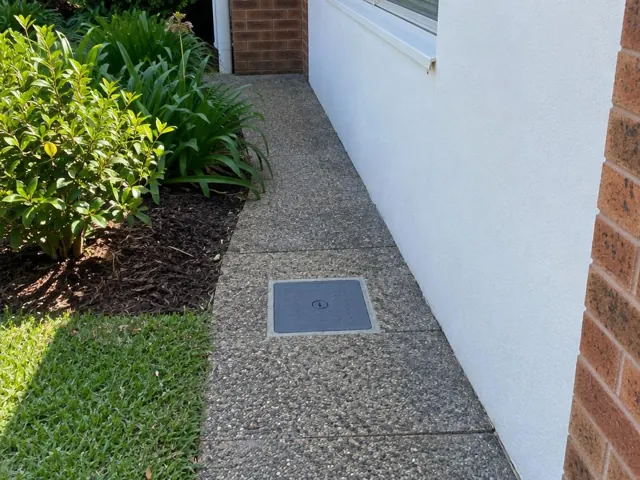 Flush inspection cover in a paved side path beside a house wall in Seven Hills