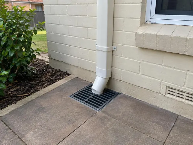 Stormwater grate beside a cream brick house wall and downpipe on paved ground in Seven Hills