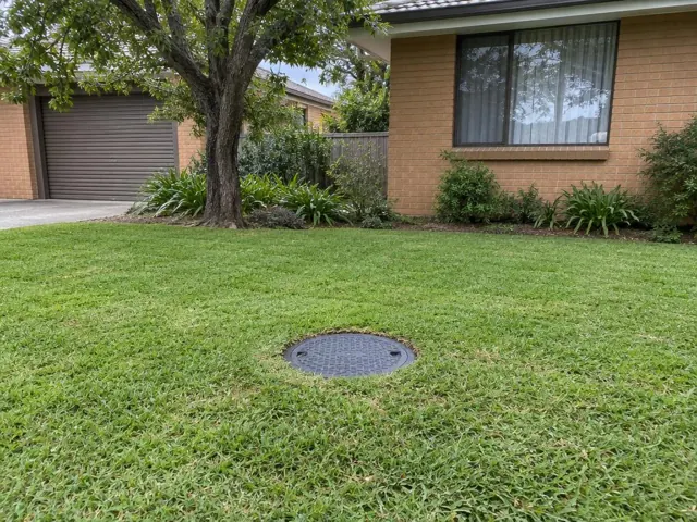 Lawn with a round capped sewer access cover beside a brick house in Toongabbie
